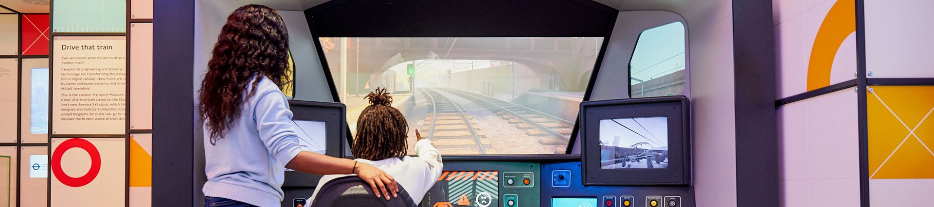 A young black girl sits in front of a train simulator with a young black woman standing next to her