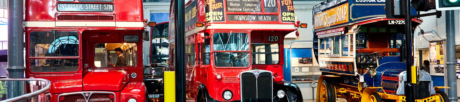 Two red Routemaster buses and a blue and yellow Leyland bus at London Transport Museum. In the foreground, the bottom of traffic light showing a green light is visible