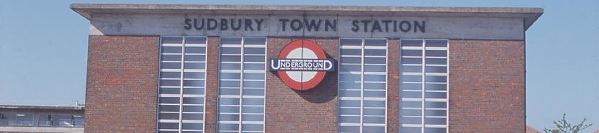 The outside of Sudbury Town station. This was designed by Charles Holden. It is rectangular brick building and with four vertical glass windows across the front. 