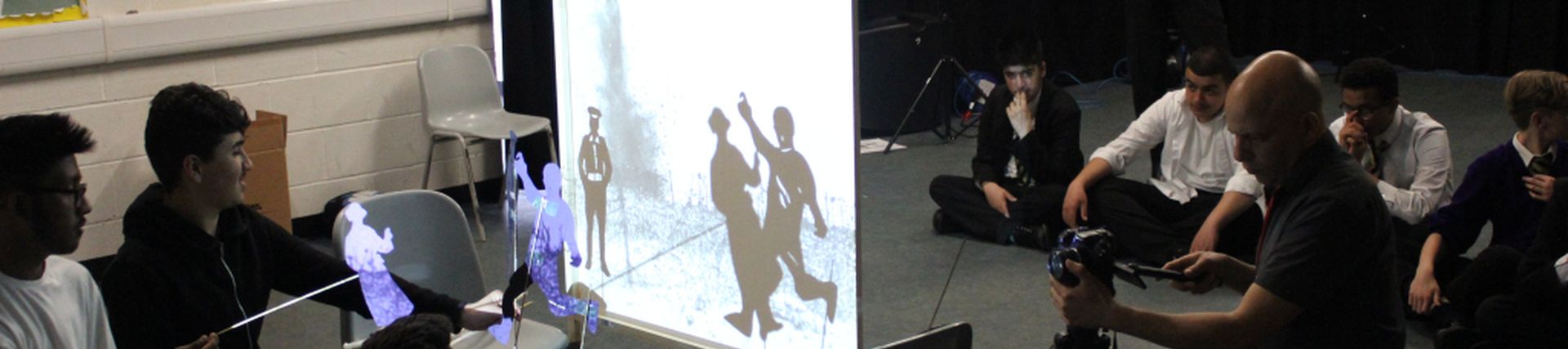 A group of people with paper cut outs of people held in front of a lit screen to portray a scene, for the camera