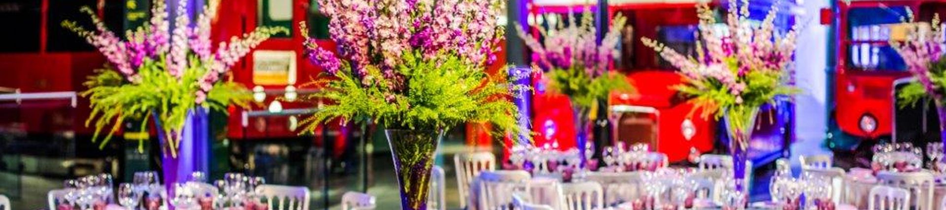 Elaborate flower displays on tables set with purple glasses in a museum in front of red London buses 